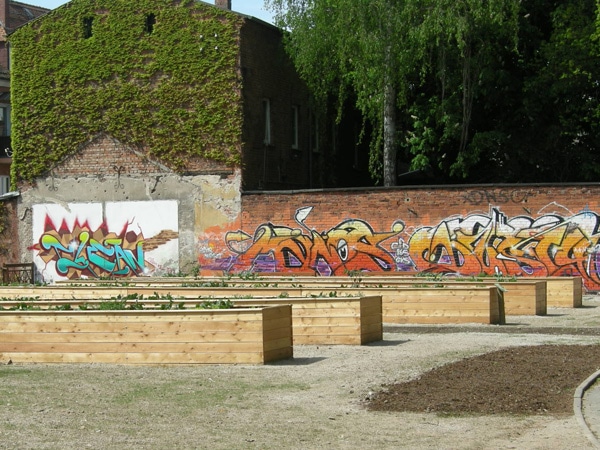 5 langgezogene rechteckige Hochbeete aus Holz, im Hintergrund Backsteinwand mit gelbrotem Graffitischriftzug