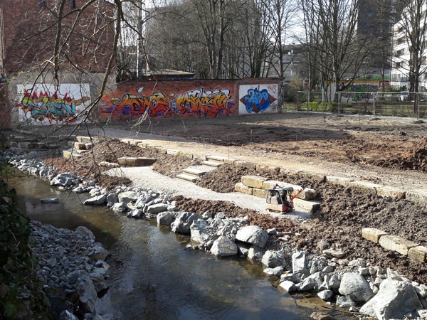 Neu gestaltete Uferböschung zum Mühlgraben mit Flussbausteinen, Schotterbänken und Sandsteinen nach Abschluss der Gartenbauarbeiten