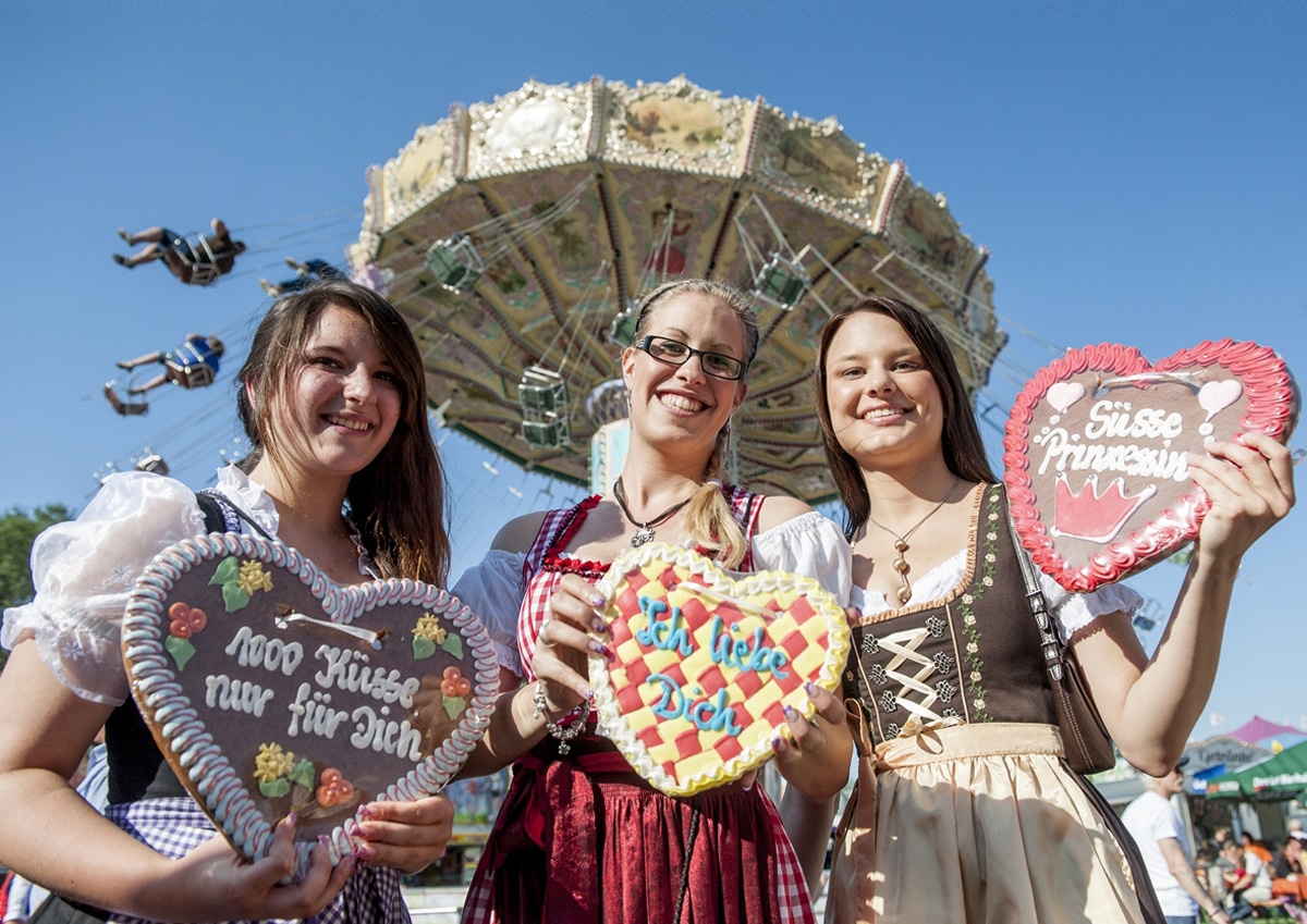 3 Frauen im Dirndl mit Lebkuchenherzen, im Hintergrund Kettenkarussell