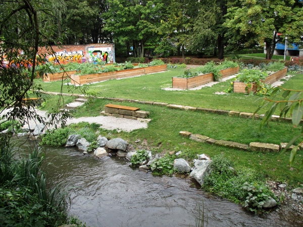 Neu gestaltete Rosenau mit bepflanzten Hochbeeten, Wiesenfläche, Sitzgelegenheiten, Mühlgraben mit Flussbausteinen und Bepflanzung