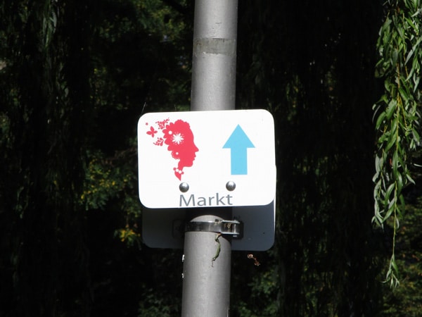 weißes Schild mit Ausschnitt des Logos LGS, blauem Richtungspfeil und Beschriftung Markt