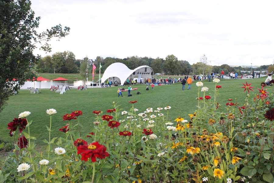 Im Vordergrund Blumenrabatte, im Hintergrund Seebühne mit weißem Foliendach