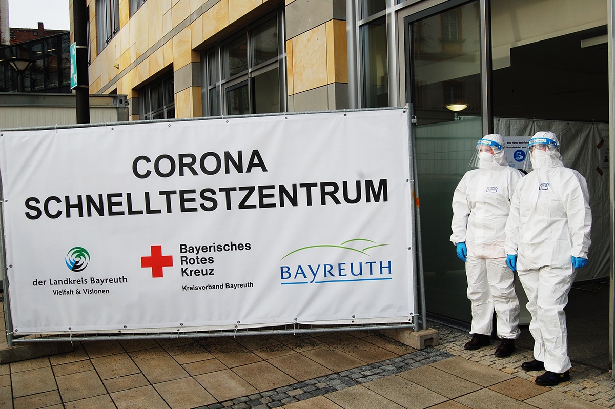 Schild und zwei Menschen in Schutzanzügen vor dem Eingang des Corona-Schnelltest-Zentrums