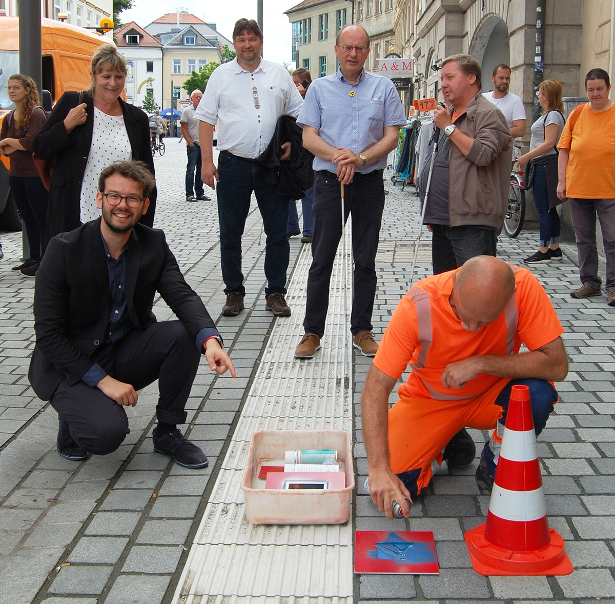 Mehrere Menschen schauen einem Mitarbeiter des Stadtbauhofs zu, wie er das Logo des Blindenleitsystems auf den Boden sprüht.