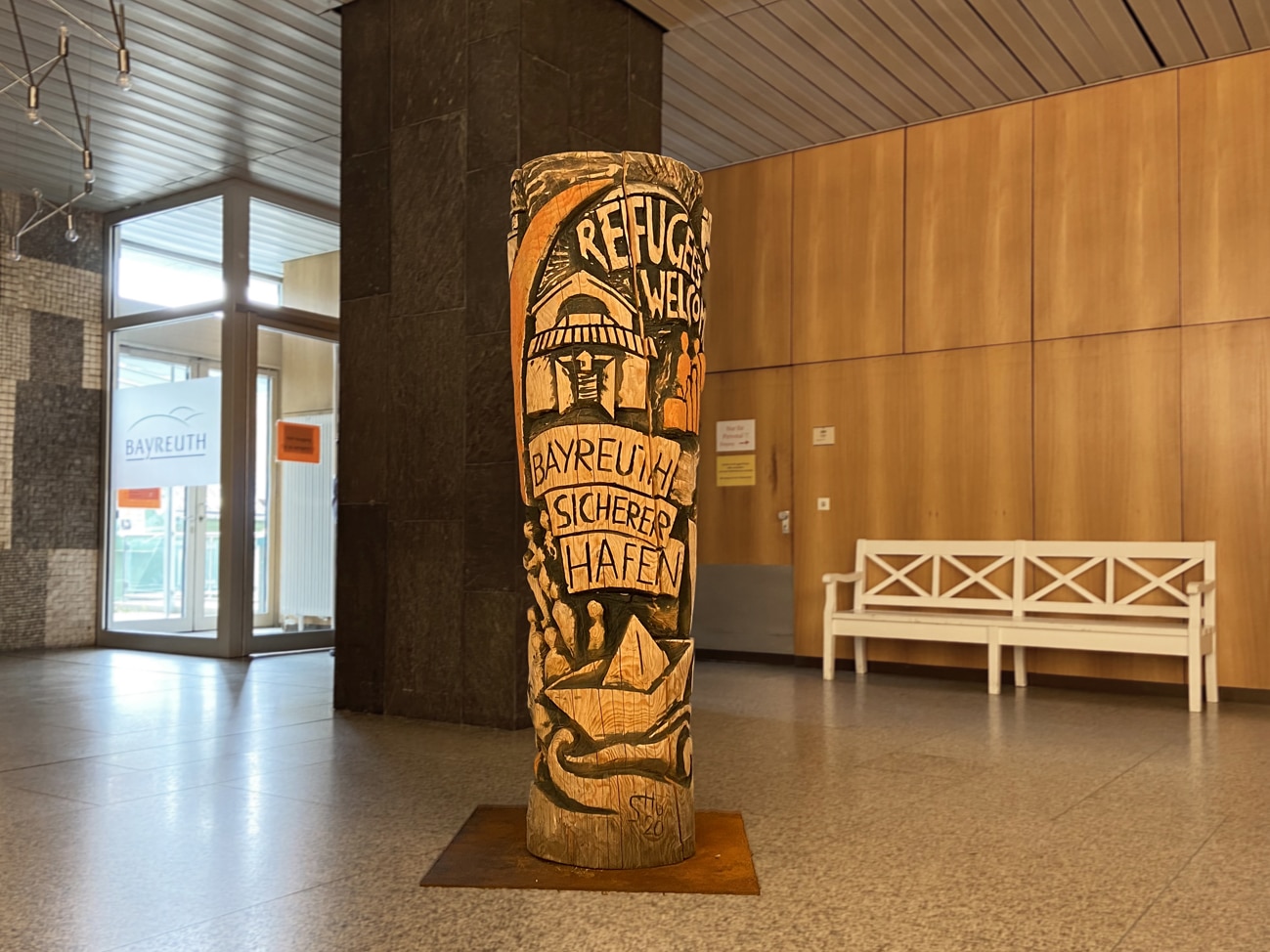 431 Skulptur.vorschau Holzskulptur vor einer weißen Sitzbank.