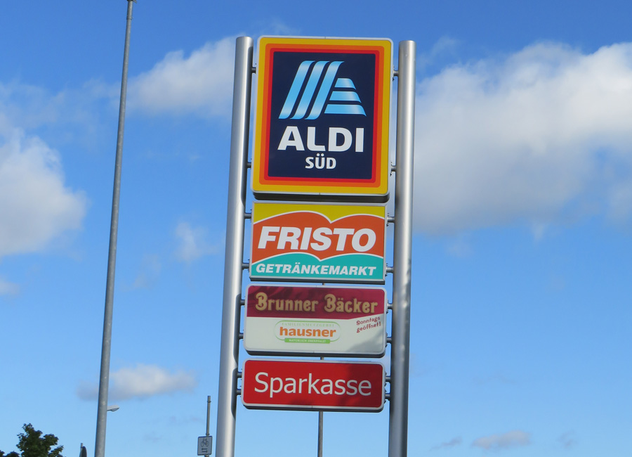 Werbetafel mit Schildern von Aldi, Fristo Getränkemarkt, Brunner Bäcker, Metzgerei Hausner und Sparkasse