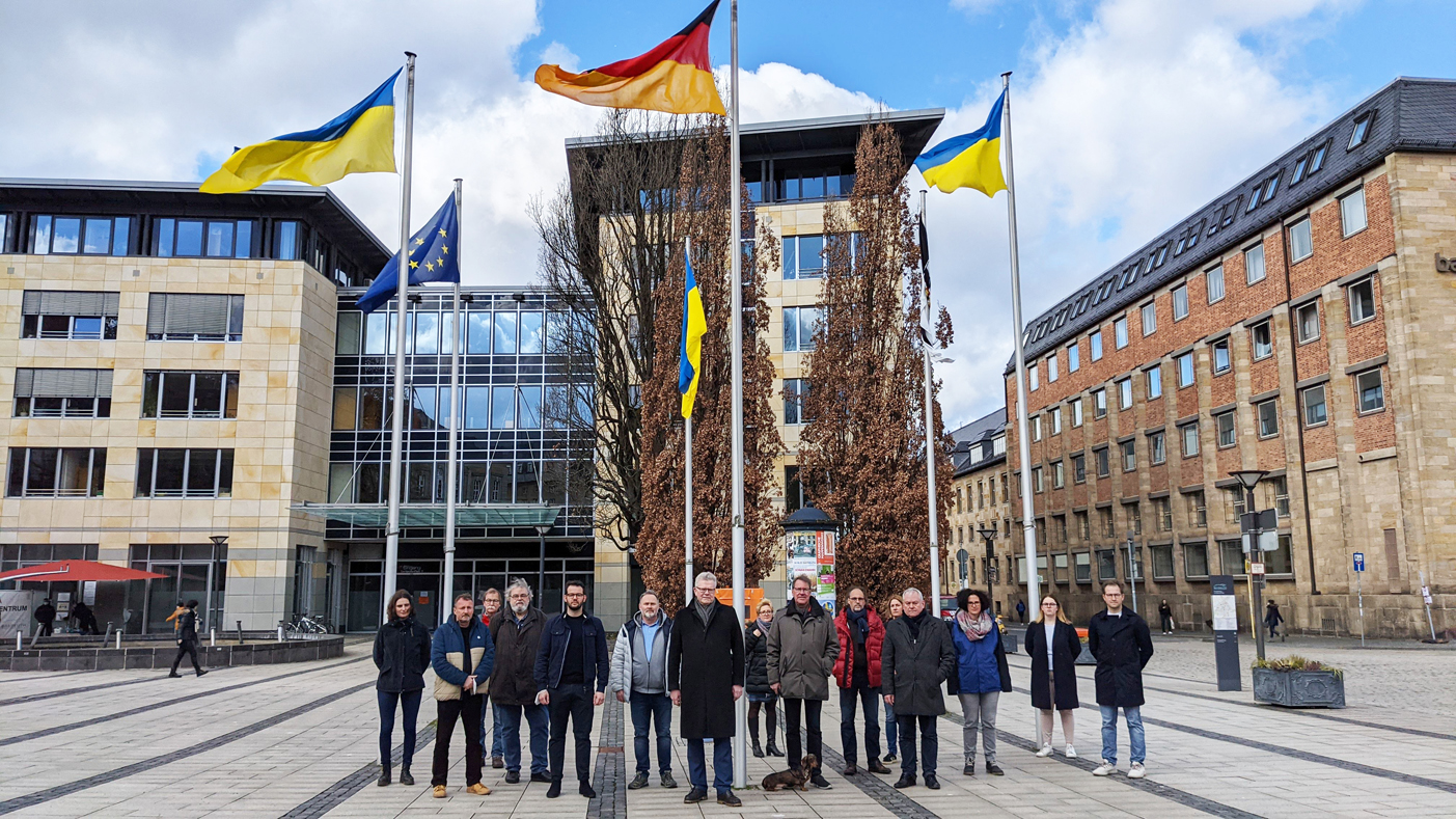 Eine Menschengruppe steht vor mehreren Fahnenmasten mit der ukrainischen Staatsflagge.