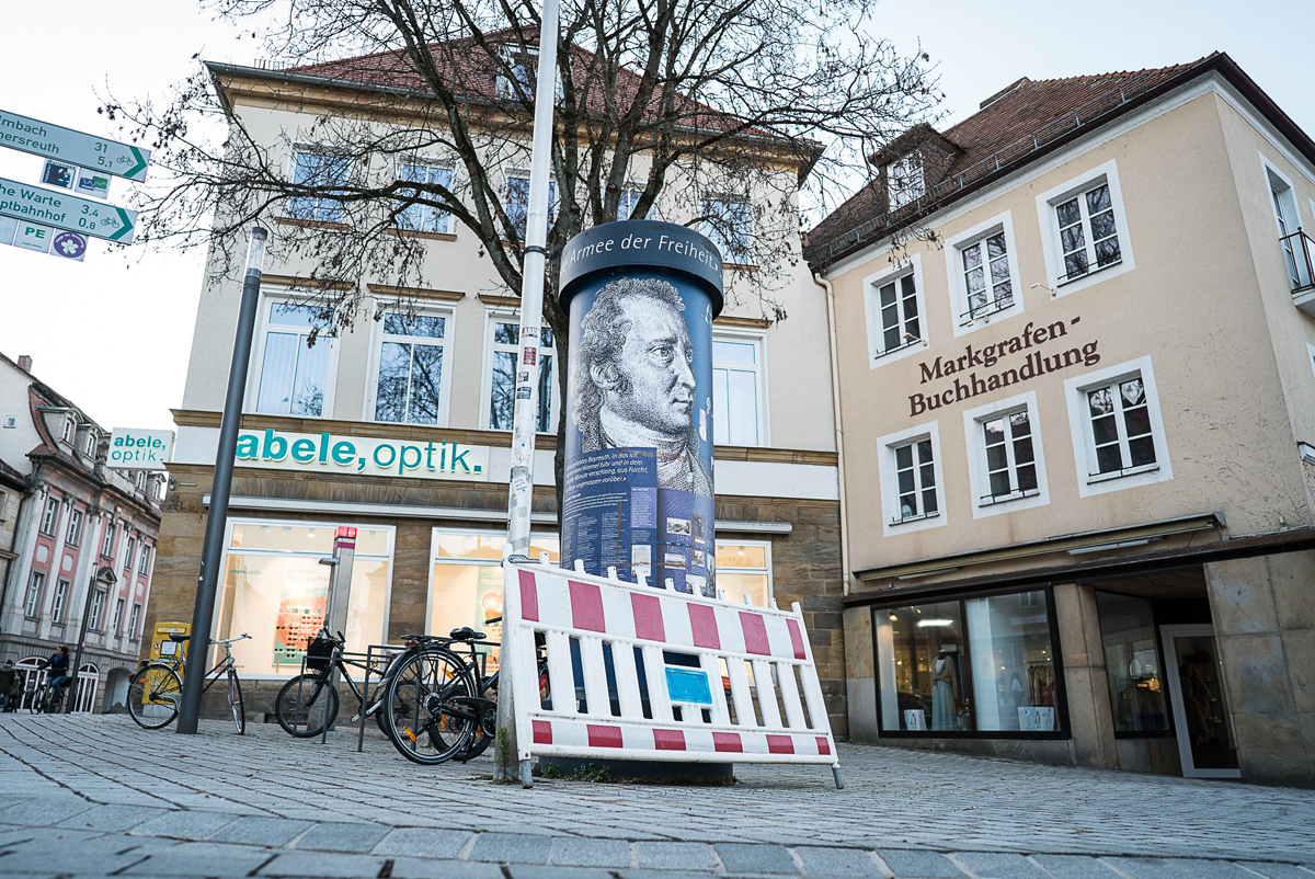 Litfaßsäule am Sternplatz mit zwei Häusern im Hintergrund.