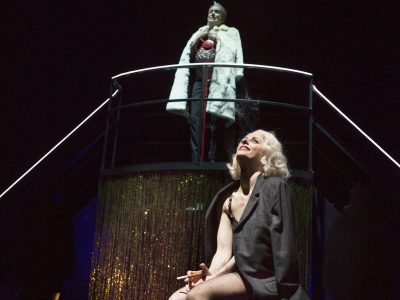 Eine blonde Frau sitzt auf einer dunklen Bühne, im Hintergrund ein Mann mit weißem Mantel.