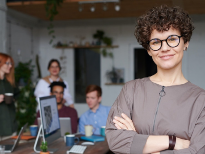 Eine junge Frau mit kurzem lockigen Haar blickt lächelnd in die Kamera. Im Hintergrund ist eine Büro-Szene zu sehen. Zwei Frauen und zwei Männer sitzen um einen Tisch herum und besprechen etwas.