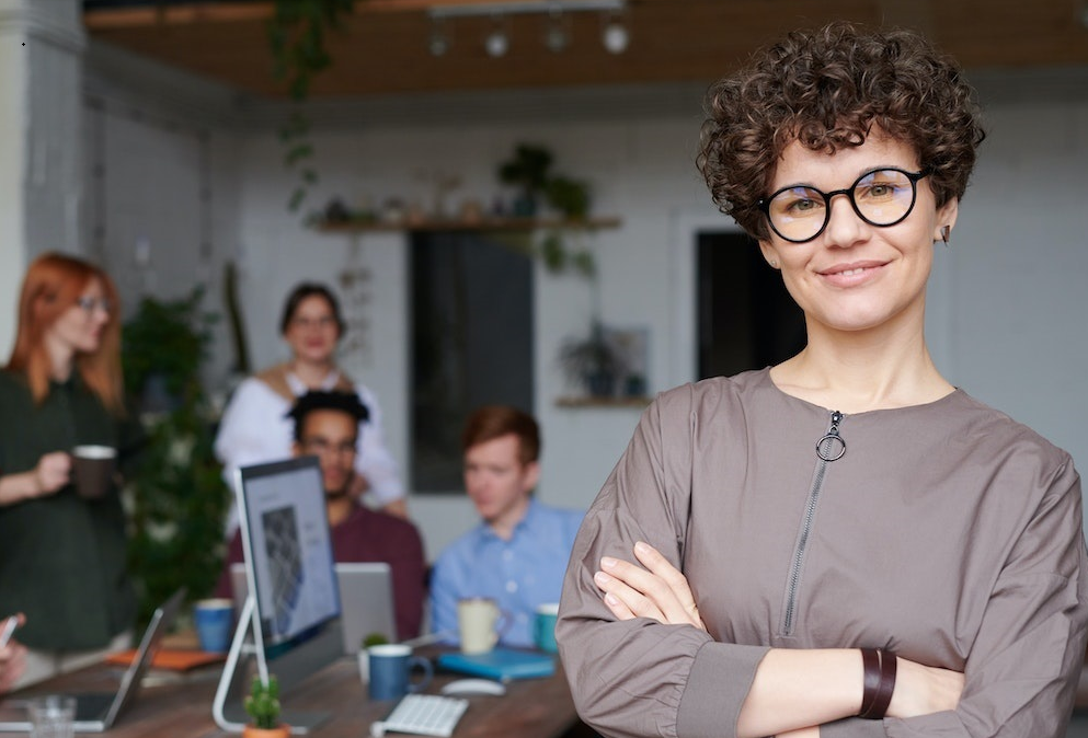 Eine junge Frau mit kurzem lockigen Haar blickt lächelnd in die Kamera. Im Hintergrund ist eine Büro-Szene zu sehen. Zwei Frauen und zwei Männer sitzen um einen Tisch herum und besprechen etwas.