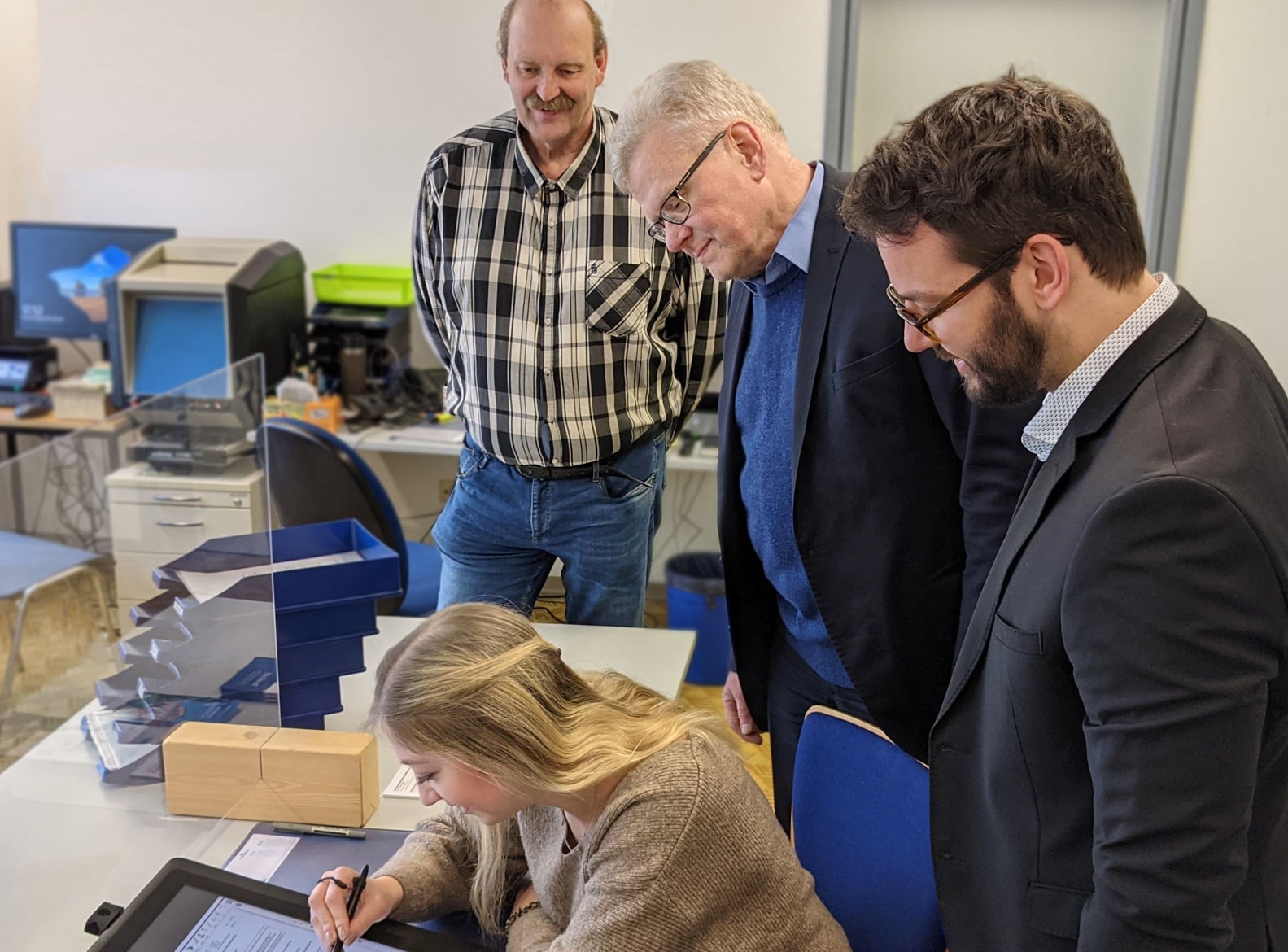 Eine junge Frau benutzt ein Signatur-Tablet. OB Ebersberger, Bürgermeister Zippel und Dienststellenleiter Ambors schauen ihr dabei über die Schulter.