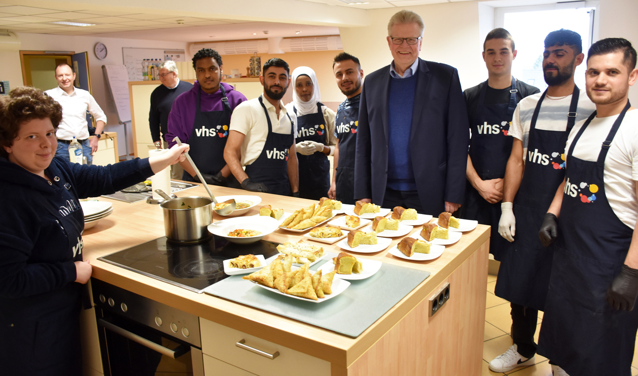 Oberbürgermeister Ebersberger steht mit Teilnehmern eines Kochkurses der Volkshochschule an einem Kochherd.