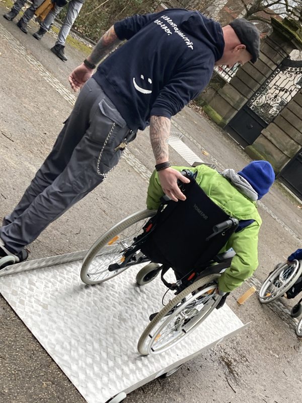 Auf dem Bild ist zu sehen, wie ein Schüler probiert mit einem Rollstuhl über eine Metallrampe zu fahren. Unterstützt durch einen Mitarbeiter des Reha-Teams.