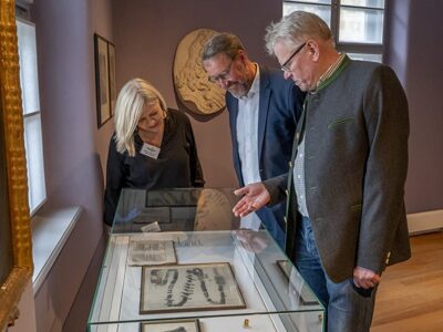 OB, Dr. Specht und Ruppert stehen vor einem Glastisch und betrachten Ausstellungsstücke.