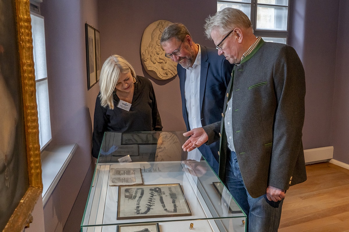 OB, Dr. Specht und Ruppert stehen vor einem Glastisch und betrachten Ausstellungsstücke.