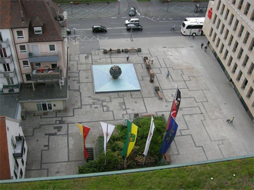 Rathausvorplatz mit unterschiedlichen, ausgebessertem Pflaster, einer Grünfläche mit Fahnen, Kugelbrunnen und wenigen Sitzbänken.
