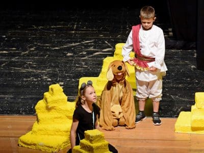 es sind drei Kinder auf einer Bühne zu sehen. Sie spielen Theater. Eines der Kinder kniet und blickt in die Ferne, neben ihr ein als Hund und ein mit einer Schärpe verkleidete weiteres Kind.