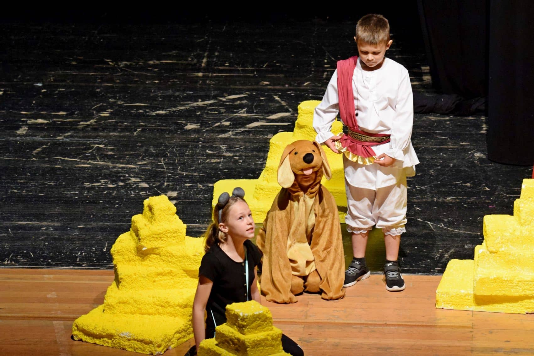 es sind drei Kinder auf einer Bühne zu sehen. Sie spielen Theater. Eines der Kinder kniet und blickt in die Ferne, neben ihr ein als Hund und ein mit einer Schärpe verkleidete weiteres Kind.