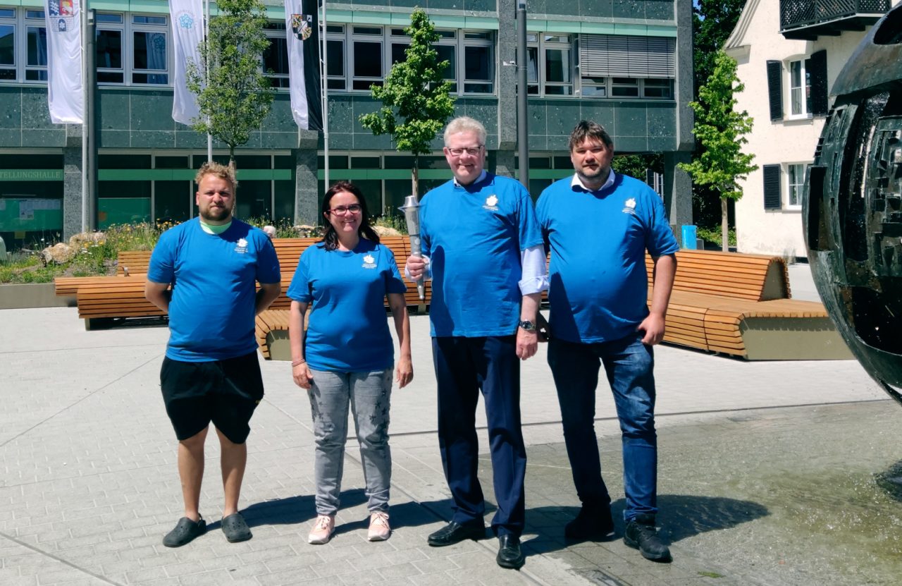Drei Männer und eine Frau in blauen T-Shirts präsentieren die Fackel für die Special Olympics World Games vor dem Rathaus.