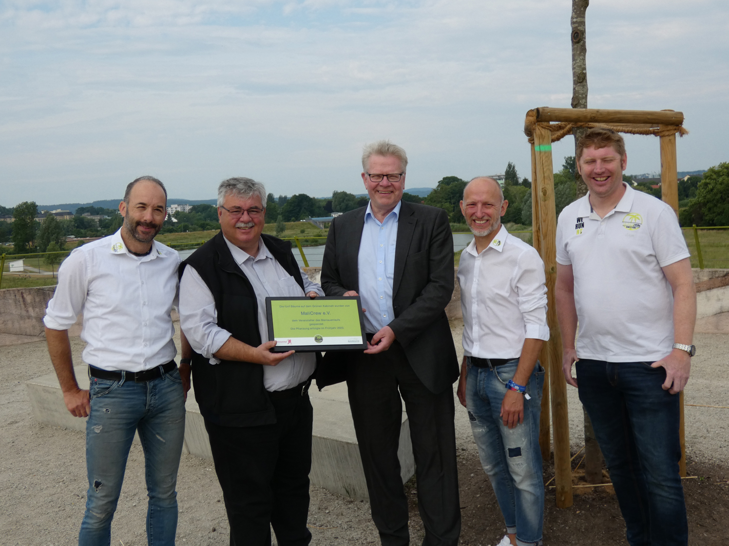Baumspende Vier Männer stehen vor einem neu gepflanzten Baum und halten einen Spenden-Scheck.