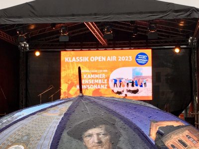 Zu sehen ist eine große LED-Leinwand auf einer Bühne. Auf ihr steht: Sparda-Bank Klassik Open Air 2023- Kammerensemble Konsonaz, Freitag 23.06.2023. Die Bühne ist perspektivisch auf gleicher Höhe wie ein Regenschirm, der das Konterfei Richard Wagners abbildet.