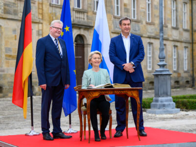 EU-Kommissionspräsidentin von der Leyen trägt sich in ein Buch ein. Neben ihr stehen Ministerpräsident Söder und Oberbürgermeister Ebersberger. | Foto: Bayerische Staatskanzlei