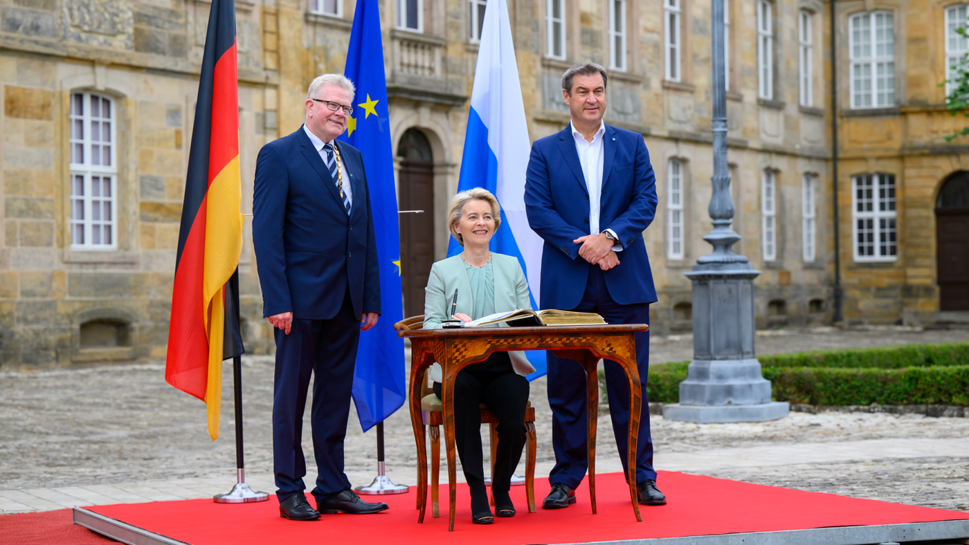 EU-Kommissionspräsidentin von der Leyen trägt sich in ein Buch ein. Neben ihr stehen Ministerpräsident Söder und Oberbürgermeister Ebersberger. | Foto: Bayerische Staatskanzlei