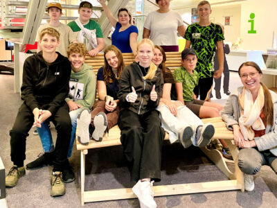 Junge Schülerinnen und Schüler sitzen mit ihren Lehrkräften auf einer Holzbank.