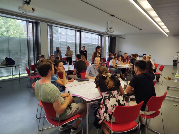 Studierende sitzen um vier Tische und erarbeiten Ideen.