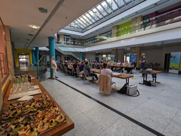 rund 30 Personen sitzen in U-Form in eiem Saal und schauen auf eine Präsentation, Im Vordergund steht ein Tisch mit Essen und Getränken