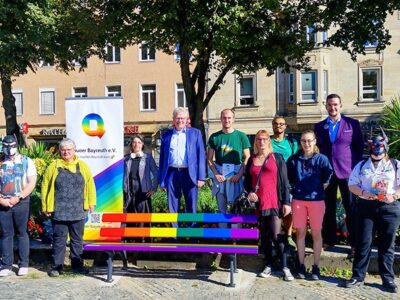 Mehrere Menschen stehen hinter der in Regenbogenfarben gestrichenen Bank