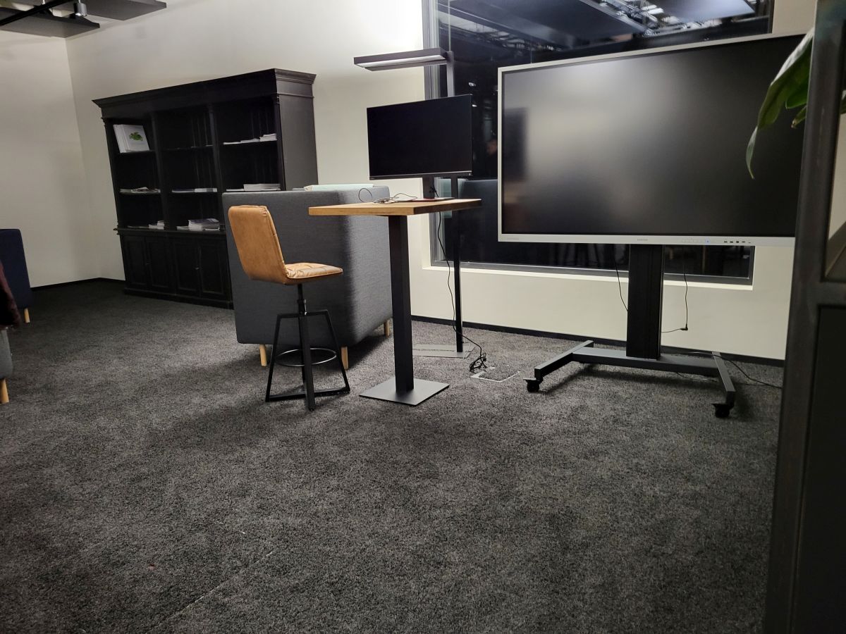 Raum für Videokonferenzen mit großem Bildschirm und einem Tisch mit Internetstation. Grauer Fußboden und helle Wände.