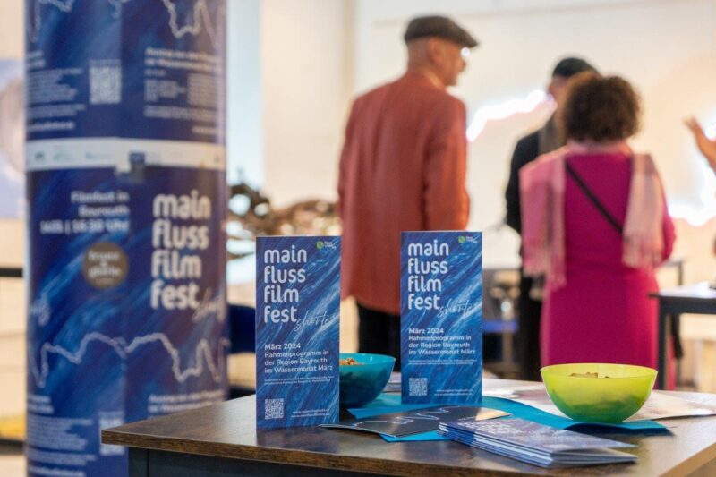 Im Vordergrund sind zwei Broschüren und Plakate der Ausstellung zu sehen im Hintergrund sind Besucher zu sehen