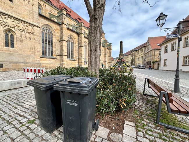 Zwei Mülltonnen stehen vor einem Baum und einem Strauch. Links ist eine große Kirche, im Hintergrund ein Osterbrunnen.