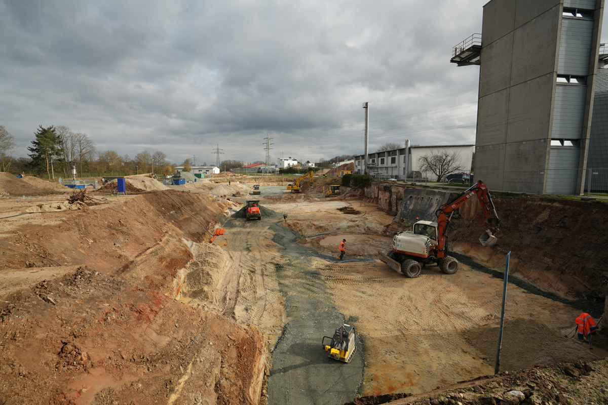 Baustelle mit Baugrube und Baumaschinen. Am rechten Bildrand ist ein Gebäude zu sehen.