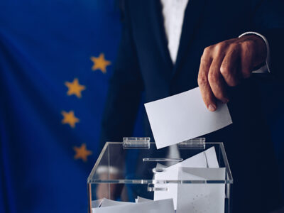 Im Hintergrund die Europäische Fahne und vorne wirft eine Hand einen Zettel in die Wahlurne