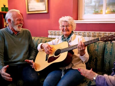 Älterer Mann und ältere Frau sitzen beieinander, Frau hat Gitarre in der Hand, es wird gesungen