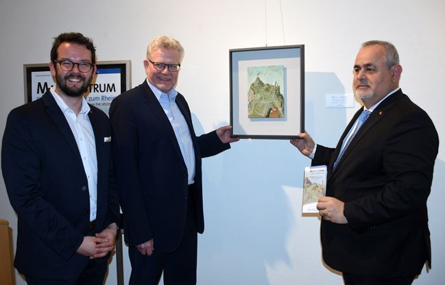 Zwei ältere Männer stehen vor einer Wand und halten ein Gemälde. Daneben steht ein dritter Mann mit Bart.