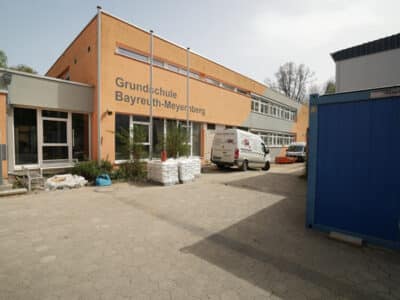 Ein Schulgebäude, vor dem Transport- beziehungsweise Handwerkerfahrzeuge parken.