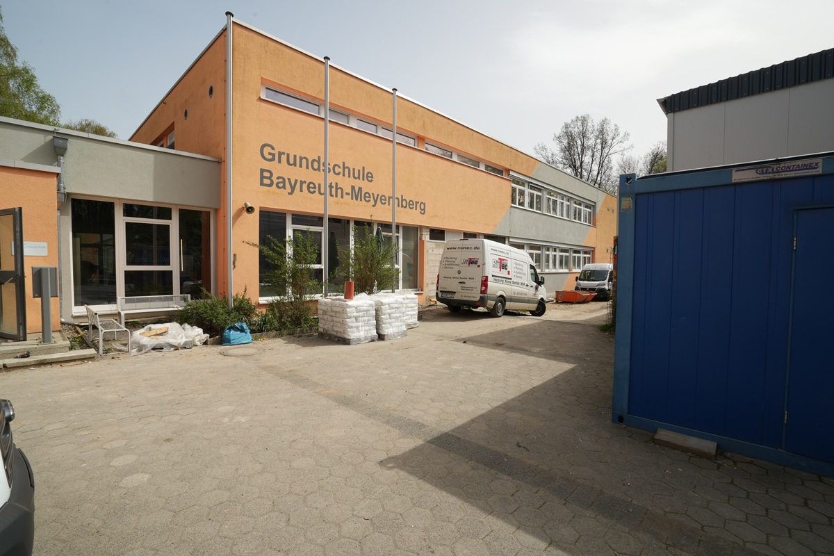 Ein Schulgebäude, vor dem Transport- beziehungsweise Handwerkerfahrzeuge parken.