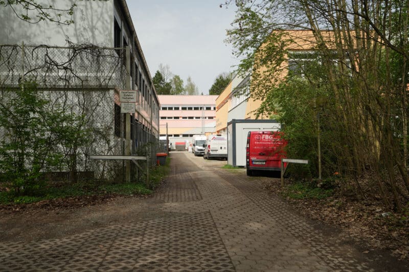Auf der linken Seite stehen Schulcontainer, rechts ist ein Schulgebäude hinter Büschen zu sehen.