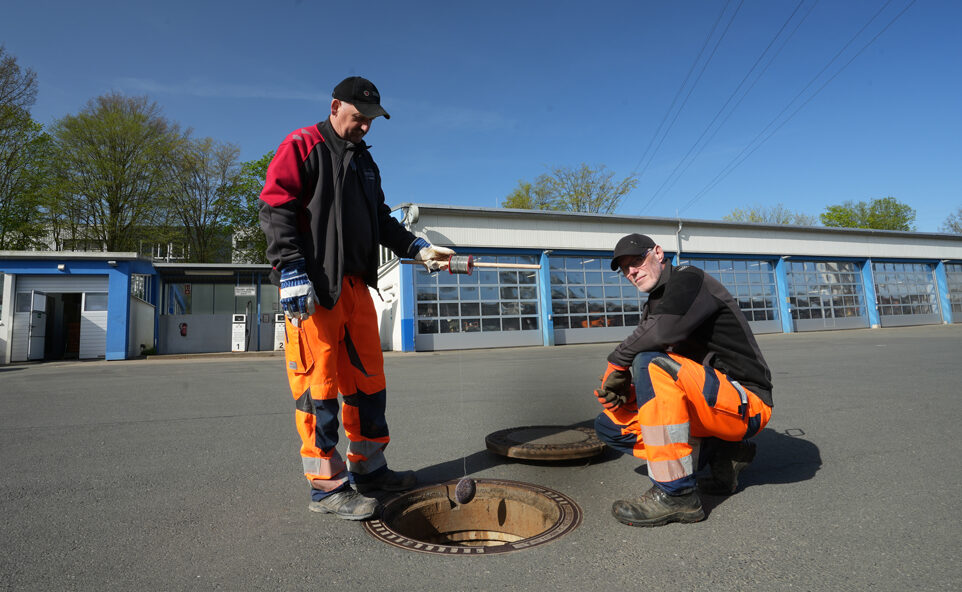 Zwei Männer des Stadtbauhofs. Einer steht, einer ist in der Hocke. Beide tragen orangene Hosen. Der stehende Mann hält einen Köder an einer Leine.