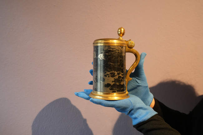 Ein Bierkrug aus Stein mit goldfarbenem Deckel, Henkel und Fuß wird von einer Hand mit blauen Handschuhen gehalten.