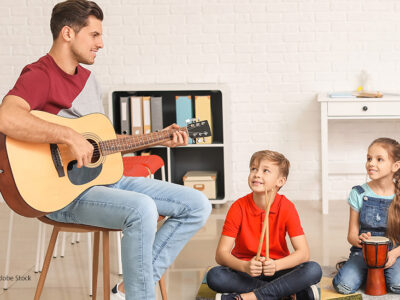Ein junger Mann spielt Gitarre. Zwei Kinder hören ihm zu. | Foto: Adobe Stock