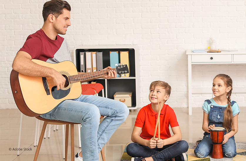 Ein junger Mann spielt Gitarre. Zwei Kinder hören ihm zu. | Foto: Adobe Stock