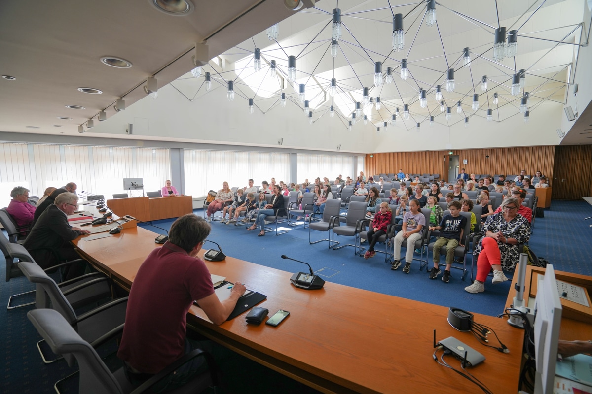 Erwachsene sprechen zu Schülern, die in einem großen Saal sitzen