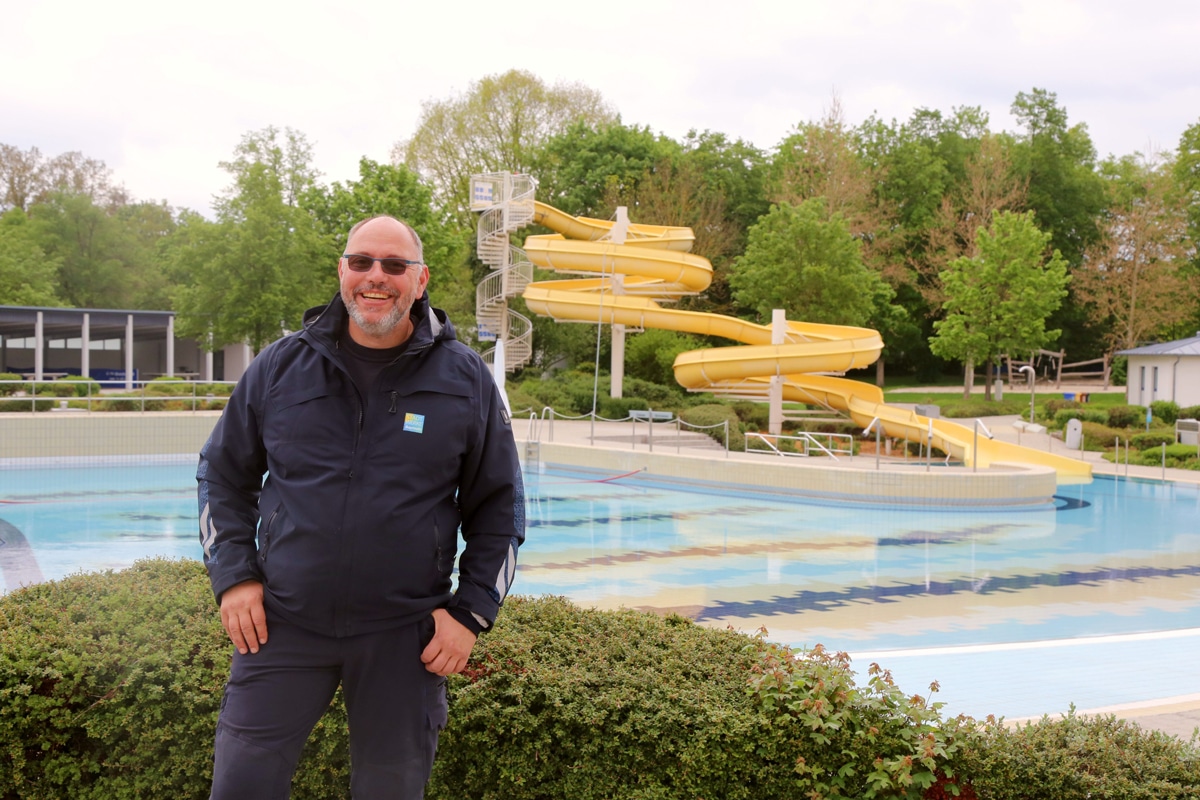 Ein Mann mit Sonnenbrille steht vor einem Freibad. Im Hintergrund ist eine Wasserrutsche zu sehen.