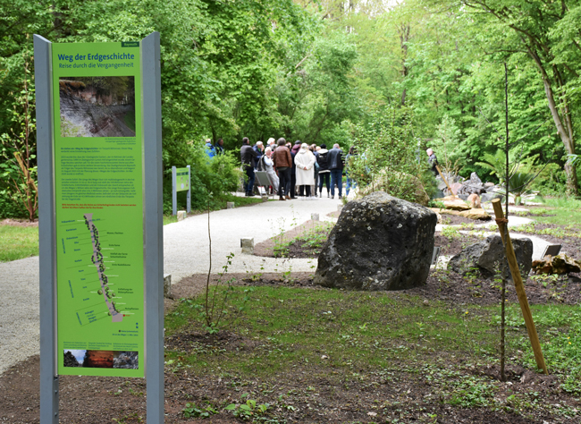 Eine Hinweistafel steht an einem Waldweg. Im Hintergrund sind Menschen zu sehen.