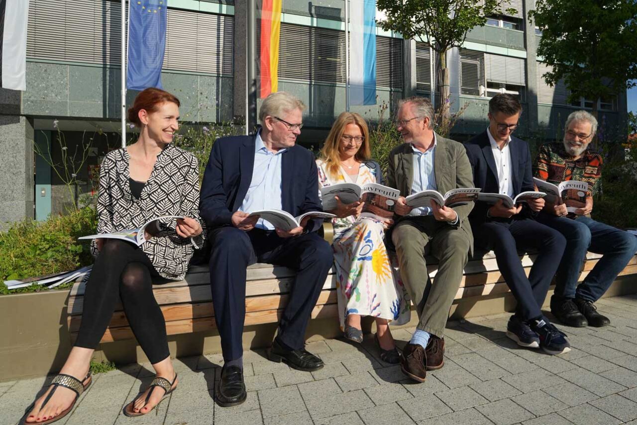Sechs Personen sitzen auf einer Bank vor dem Rathaus und blättern in einem Magazin. | Foto: Stadt Bayreuth